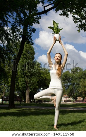 young slim woman standing in yoga  pose in park, holding green flower in pot, near big green tree