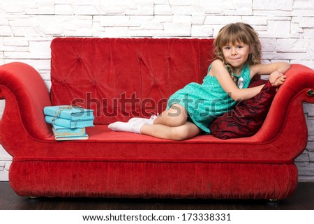 little beautiful girl in fashion dress with long blond curl learn on red cough, near learn pile of old books