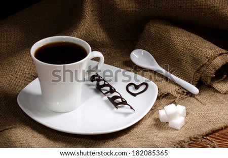 on white plate - coffee in white cup and chocolate slice and chocolate heart, near - ceramic spoon and sugar lumps on sacking, on wooden table