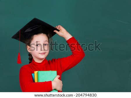 little schoolgirl holding colorful books and  trying university hat on chalkboard background