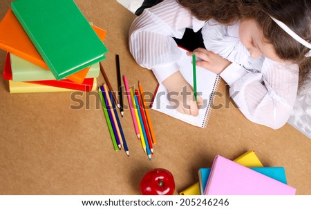 little schoolgirl writing in copybook, near lye red apple, many colorful books and pencils, top view