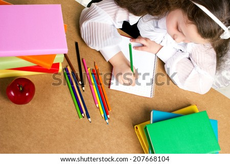 beauty little girl  in school form diligently writing in copybook, near -  red apple, many colorful books and pencils, top view
