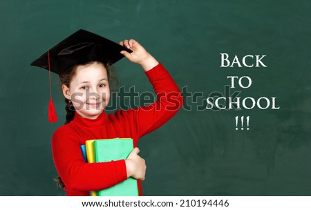 little schoolgirl holding colorful books and  trying university hat on chalkboard background