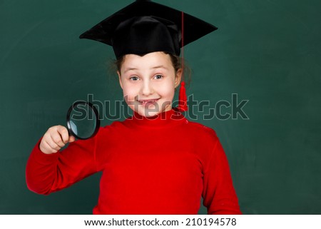 little joyful schoolgirl holding magnifying glass and  trying university hat on chalkboard background