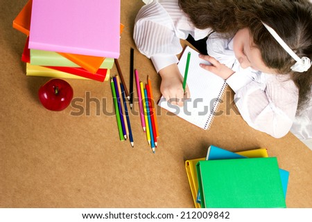 beauty preschooler girl  in white and black uniform diligently writing in copybook, near -  red apple, many colorful books and pencils, top view