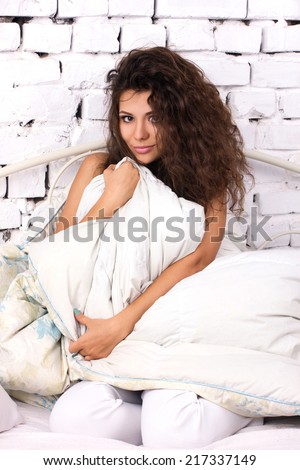 beautiful brunette young woman with long dishevelled curls sitting in the bed under duvet on white bricky wall background