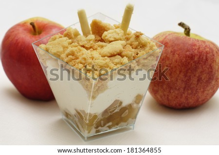 A layered dessert in a plastic serving dish, with apples behind it. The dessert consists of apples, chantilly cream with vanilla specks, and a crumbly streusel topping.