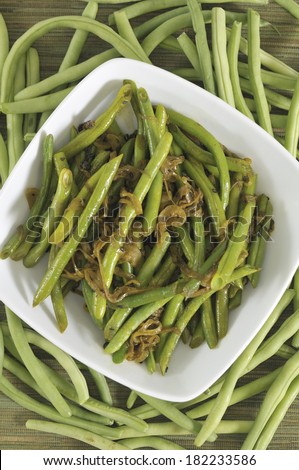 An overhead view of a vegetable dish of sauteed green beans, onions and curry spices, surrounded by raw green beans