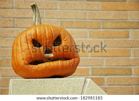 A pumpkin carved to show an angry expression sitting on a white stone column against a light brick wall.  The mouth has dried and shrunken to create a grimace expression