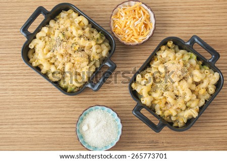Two pans of macaroni and cheese with broccoli, accompanied by two side dishes of parmesan and cheddar cheese to add