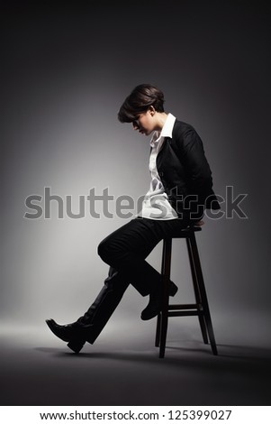 cool young woman in suit sitting on stool and looking down on grey background