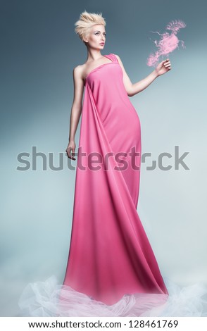 beautiful young model holding a peacock made of smoke wearing long pink dress and posing on blue background