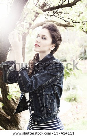 beautiful young lady with a blue jean jacket touching the leaves of a tree outdoors