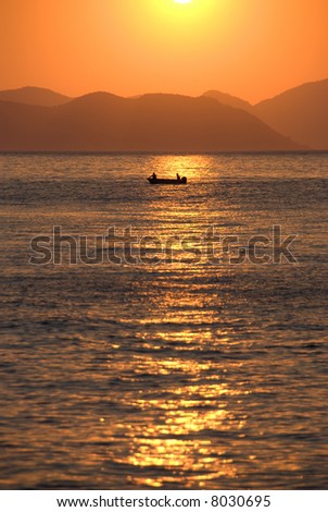 Sun sets over the Mediterranean sea, with the silhouette of a fishing boat in the distance.
