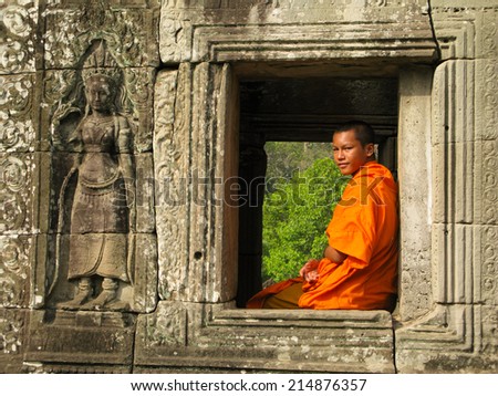 Buddhist monk sitting, framed by ancient stone window at Angkor Wat temple complex, Cambodia (1. September 2014). Clad in orange robe, young, looking at the camera, green tree foliage background.