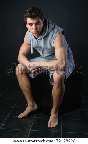 Muscle young boy sits in grey clothes on a black cube in the dark
