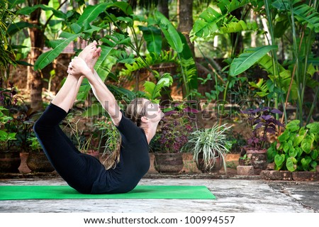 Yoga Dhanurasana bow pose by woman in black cloth in the garden with palms, banana trees and plants in the pots