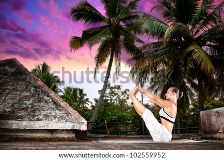Yoga naukasana boat pose by woman in white cloth on the roof at palm trees and sunset background in Varkala, Kerala, India
