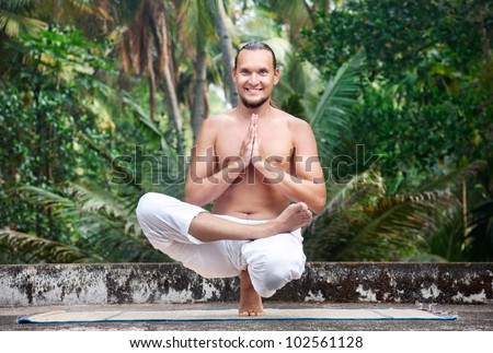Yoga Ardha Baddha Padma Padangusthasana balancing on toes by man with beard in white trousers on the roof at palms background
