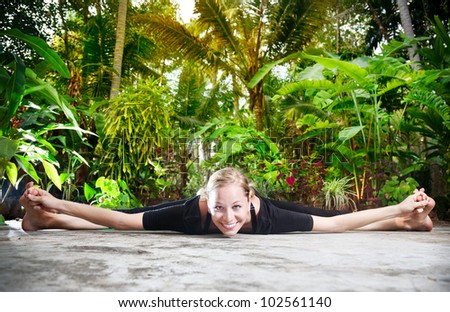 Yoga upavistha konaasana pose by woman in black cloth in the garden with palms, banana trees and plants in the pots