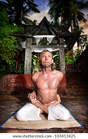 Yoga meditation in baddha padmasana pose by man in white trousers at stone temple background in tropical forest