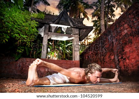 Yoga upavistha konasana pose by man in white trousers near stone temple at sunset background in tropical forest