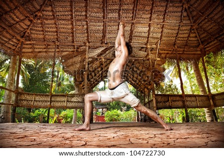 Yoga virabhadrasana I warrior pose by fit man in white trousers on the drought earth in yoga shala, Varkala, Kerala, India