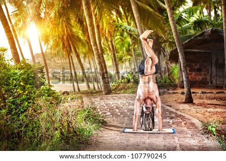 Yoga handstand pose with legs in garudasana by fit man with dreadlocks on the beach near the fishermen hut in Varkala, Kerala, India