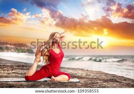 Yoga raja kapotasana pigeon pose by young woman with long hair in red cloth on the beach near the ocean at dramatic sunset background