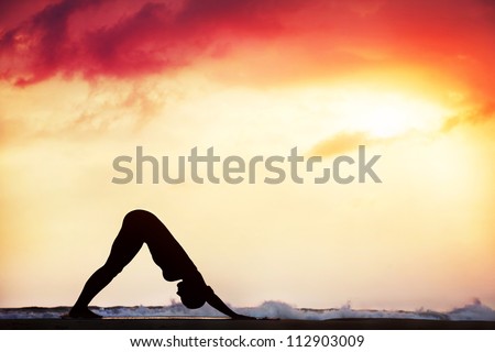 Step of surya namaskar, downward facing dog pose by beautiful woman on the beach near the ocean at dramatic sunset background
