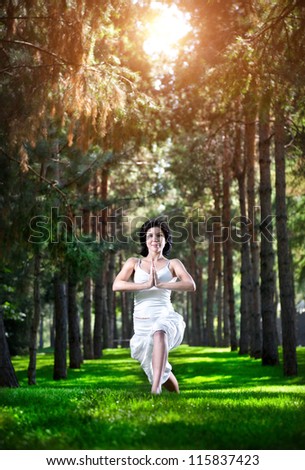 Yoga virabhadrasana I warrior pose by woman in white costume on green grass in the park around pine trees