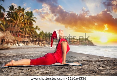 Yoga cobra pose by young woman in red costume and red christmas hat on the beach near the ocean at sunset background in India