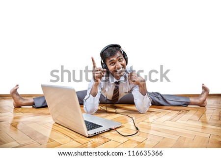 Indian businessman listening the music with headphones and doing yoga near the laptop in the office at white background. Free space for your text