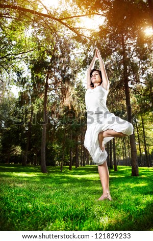 Yoga tree pose by woman in white costume on green grass in the park around pine trees