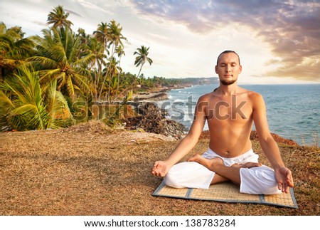 Yoga meditation in lotus pose by man in white trousers on the cliff near the ocean in Kerala, India