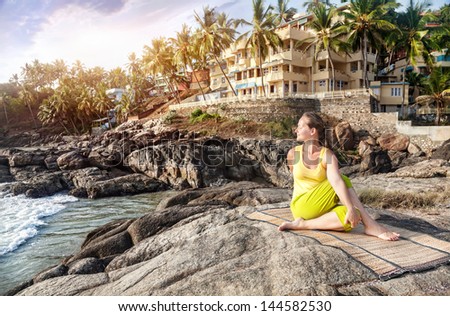 Yoga by woman in yellow costume on the stone nearby ocean and tropical resort in Kovalam, Kerala, India