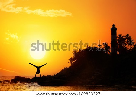 Man doing Yoga handstand on the rocks near lighthouse at sunset sky