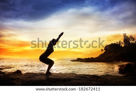 Woman doing Yoga Utkatasana pose in silhouette on the stones near lighthouse at sunset sky in Kovalam, Kerala, India