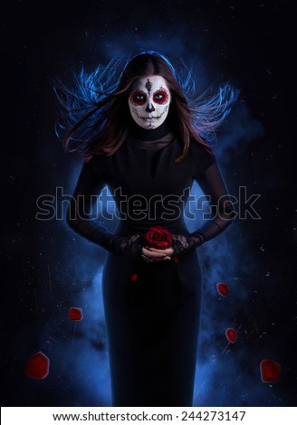 Woman in black dress with sugar skull makeup holding red rose with falling petals at dark background