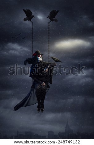 Woman with sugar skull makeup holding black crow and flying above the city at dark overcast sky with snowfall