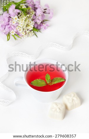 Cup of hibiscus tea with mint leaves, white chocolate candies and flower bouquet on the white table at breakfast time