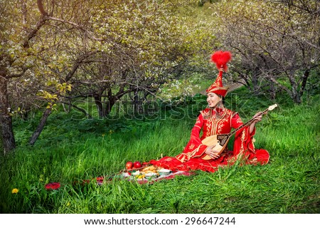 Kazakh woman in red costume playing dombra on the green grass in the apple garden of Almaty, Kazakhstan, Central Asia