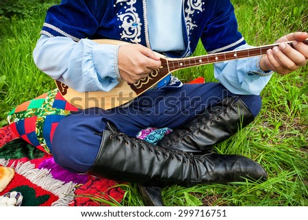 Man in traditional Kazakh costume playing dombra instrument on the green grass in apple garden of Almaty, Kazakhstan, Central Asia