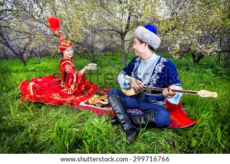 Kazakh woman with tea and man playing dombra instrument on the green grass in apple garden of Almaty, Kazakhstan, Central Asia