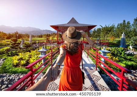 Woman in orange dress and hat holding man by hand going to Japanese Garden with pagoda