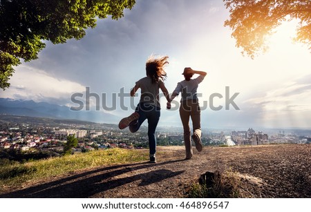 Two friends holding hands and running to the beautiful view of city panorama in the mountains at rainy dramatic sunset