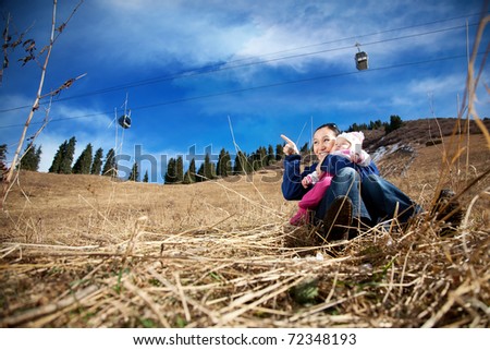 Asian mother hugging her baby and pointing at free space for text in the mountains. Ropeway with funicular and dramatic sky at the background