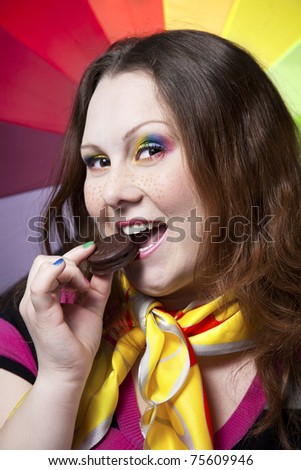 Beautiful woman with freckles, creative rainbow make-up and nails biting chocolate cookie at rainbow background