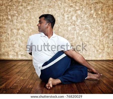 Handsome Indian man in white shirt doing ardha matsiendrasana twist pose indoors on wooden floor at grunge background