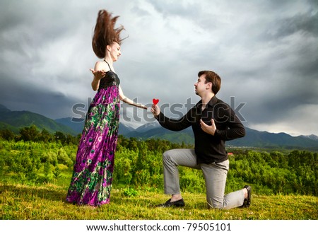 Crazy Woman in peacock dress with hair in the air and Young man giving red heart to her at mountain background with dramatic sky